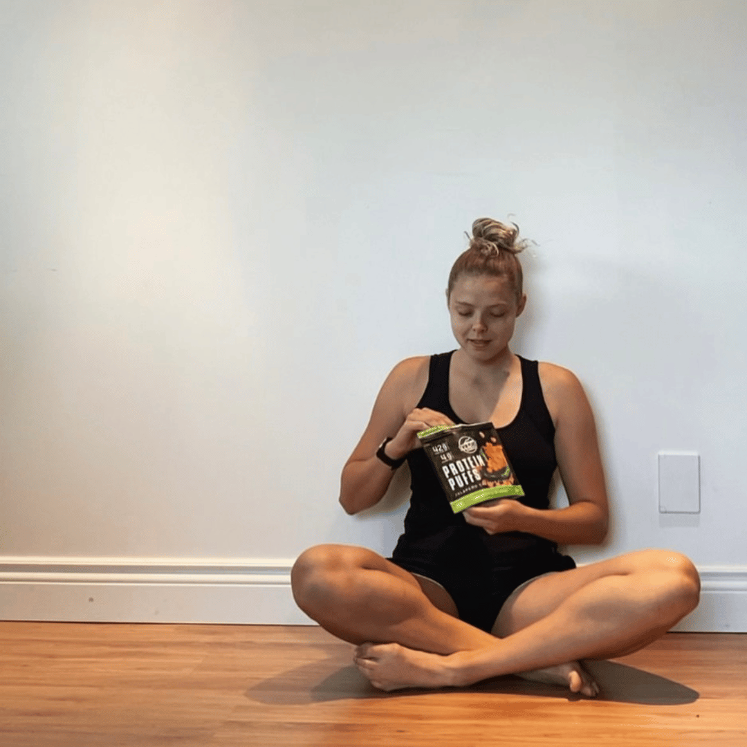 Girl Sitting Criss Cross Legs on Floor Eating Jalapeno Cheddar Protein Puffs Out of Bag